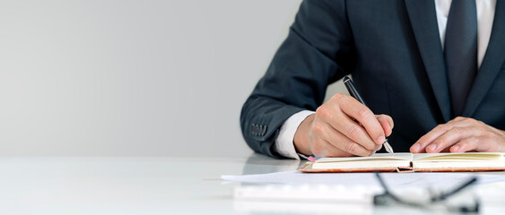 Young businessman in suit writing business plan at desk in modern office room with copy space.