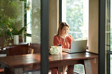 Wall Mural - Young woman sitting at cafe table, drinking fruit cocktail and answering e-mails of coworkers or clients of studying online