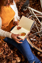 Woman Keeping Leaf In Book Sitting By Bicycle At Autumn Forest