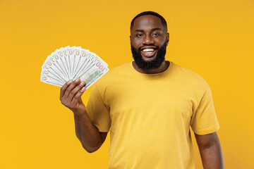 Wall Mural - Young smiling happy rich black man 20s wearing bright casual t-shirt holding fan of cash money in dollar banknotes isolated on plain yellow color background studio portrait. People lifestyle concept.