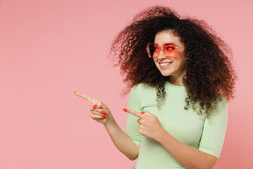 Wall Mural - Fun young curly latin woman 20s years old wear mint t-shirt sunglasses pointing fingers hands aside on workspace area copy space mock up isolated on plain pastel light pink background studio portrait