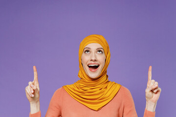 Surprised happy young arabian asian muslim woman in abaya hijab yellow pointing overhead on workspace area copy space mock up clothes isolated on plain pastel light violet background studio portrait.