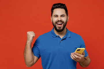 Wall Mural - Young smiling fun cool happy excited man 20s in basic blue t-shirt hold in hand use mobile cell phone browsing do winner gesture isolated on plain orange background studio. People lifestyle concept.