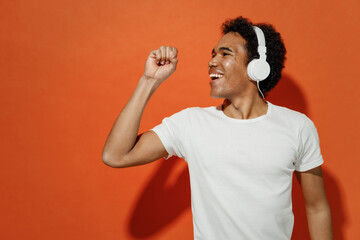 Young black curly man 20s wears white t-shirt listen music in headphones sing song fooling around have fun gesticulating hands relax enjoy isolated on plain pastel orange background studio portrait.