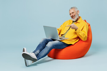 Full body smiling cool elderly gray-haired mustache bearded man 50s wear yellow shirt sit in bag chair hold use work point on laptop pc computer isolated on plain pastel light blue background studio