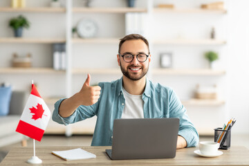 Poster - Happy young guy sitting at table with flag of Canada, using laptop computer, showing thumb up gesture at home