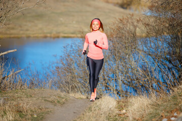 Wall Mural - Young woman jogging in the park near the pond in the cold sunny morning. Sporty girl running in countryside