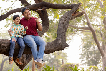 Cheerful father and son sitting on tree branch while admiring nature at park
