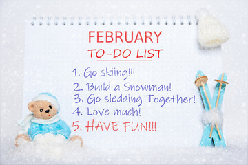 February to-do list. Notepad with to-do list and a toy Teddy bear in blue clothes, blue skis, a white hat on white snow