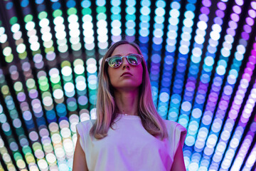 Teen hipster girl in stylish glasses standing on blue tunnel with neon light wall background, female teenager fashion model pretty young woman looking at night club city light glow