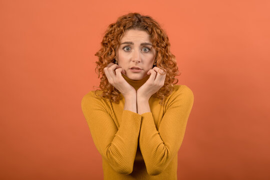 Portrait of a frightened and shocked redhead Caucasian girl in orange jumper isolated on orange studio background.