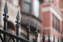 Closeup Of Repeating Ornate Iron Fence With Selective Focus On One Spire, With Blurred Brown Townhouses In The Background