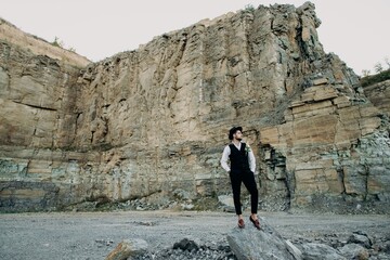 Handsome brunette man with a beard in a suit who is stacked with a white shirt, black trousers and a black vest, stands in a black hat and with flowers on a stone, near mountains and stones