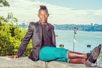 Wall Mural - Man Traveling. Dressing in a black blazer,  green pants, a young black guy with mohawk hair is sitting on a rocky stone by a river, stretching his legs, relaxing.  A USA flag in the background..