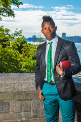 Wall Mural - Modern Businessman. Wearing a black blazer, green pants, green neck tie, a hand holding a red book under arm, a young black guy with mohawk haircut is standing by a river, looking at you..