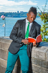 Wall Mural - Dressing in a white undershirt, a black blazer, green pants, a green tie, holding a red book,  a young black guy with mohawk haircut is standing by a river,  reading outside..