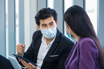 Millennial Asian male professional successful businessman manager in formal suit wearing safety face mask sit talking discussing via tablet and laptop computer with businesswoman colleague in office