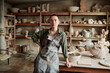 Portrait of professional sculptor in apron looking at camera while working in workshop with ceramic sculptures