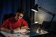 Young Asian man wearing a red T-shirt is writing papers on a desk in the dark late at night at home.