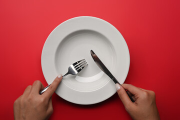 Woman holding fork and knife in hands with empty plate over red background.