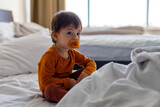 Fototapeta  - A one-year-old child with a pacifier in his mouth is sitting on the bed in the bedroom.