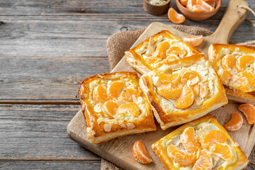 Wall Mural - Mini puff pie with cheese cream, almonds, and tangerines on a wooden serving board on the kitchen table. Delicious layered square with citrus fruits, dorblue or ricotta, nuts on a culinary background