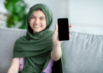 Wall Mural - Muslim teenage girl in hijab showing cellphone with blank screen at home, mockup for mobile app or website design