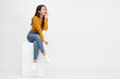 © comzeal - Portrait of young Asian woman sitting on white box and keeps hand near mouth and whispers secret isolated over white background, Spreads rumors concept