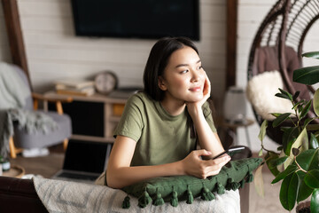 Dreamy asian girl spend time at home, holding smartphone and sitting on couch, smiling while looking at window