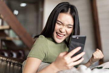 Smiling asian woman looking satisfied, making fist pump and reading mobile phone, sitting at home and triumphing