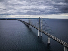 Aerial View Of Sailboat Sailing Under Oresund Bridge, On A Dramatic Windy And Cloudy Day, Denmark.