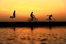 Silhouette Of Person Riding Witches Broom And Person Riding A Bicycle And Another Person Moving A Wheel Near Body Of Water During Sunset