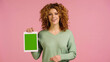 happy redhead woman in green jumper holding digital tablet with green screen isolated on pink