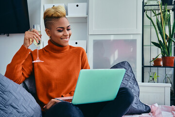 Smiling young woman drinking from champagne flute while video calling through laptop at home