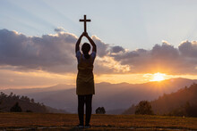 Silhouette Of Young Girl Praying To The GOD While Holding A Crucifix Symbol With Bright Sunbeam On The Mountain