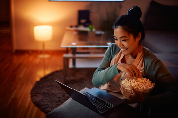 Happy Asian woman eats popcorn while watching movie on laptop at night at home.