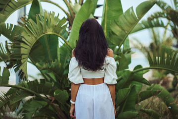 Rear view of gorgeous brunette woman in summer beach outfit relaxing outdoors against palm trees on the background. A fashionable romantic young adult lady wearing a trendy white blouse, and skirt