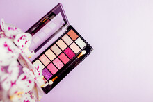 Palette Of New Colourful Eyeshadows, Pigments, Glitter With Orchid Flowers. Make-up Products On Purple Background