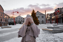 The Girl Covered Her Eyes With Her Hands. Protect Yourself From The Winter Wind Outside. A Woman Waiting For A Surprise Near A Christmas Tree At A Festive Fair In Europe