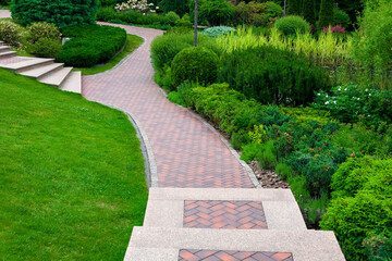 Wall Mural - stone steps descent to a path from paving slabs in a park on a slope with ornamental plants, evergreen bushes and trees, a backyard landscape a place to be at peace.