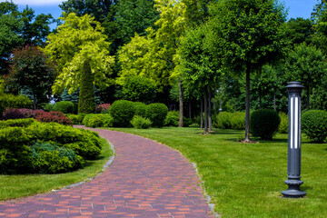 Wall Mural - lantern iron ground garden lighting of park curved path paved with stone tiles in backyard among plants, evergreen bushes and foliage trees surrounded by green lawn on sunny summer day, nobody.