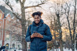 Young smiling African man wearing jacket and backpackholding his smartphone standing on the street of city. Tourism, vacation in Europe, communications. Male look at camera