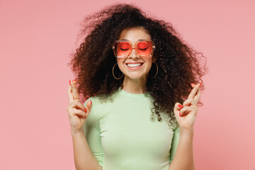 Canvas Print - Fun young curly latin woman 20s wears mint t-shirt sunglasses waiting for special moment keeping fingers crossed making wish eyes closed isolated on plain pastel light pink background studio portrait.