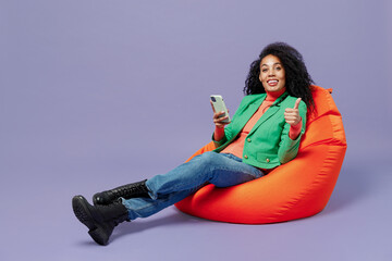 Wall Mural - Full size young black curly woman 20s wears casual clothes sit in bag chair hold in hand use mobile cell phone show thumb up like gesture isolated plain pastel light violet background studio portrait.