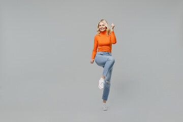 Full body elderly overjoyed excited happy blonde woman 50s in orange turtleneck do winner gesture raise up leg clench fist isolated on plain grey background studio portrait. People lifestyle concept.