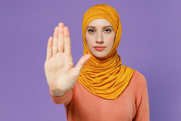 Wall Mural - Displeased irritated dissatisfied severe young arabian asian muslim woman in abaya hijab yellow clothes showing stop gesture with palm isolated on plain pastel light violet background studio portrait.