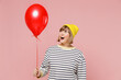 Elderly amazed woman 50s in striped shirt yellow hat celebrating birthday holiday party and hold colorful air inflated helium balloon isolated on plain pastel light pink background studio portrait