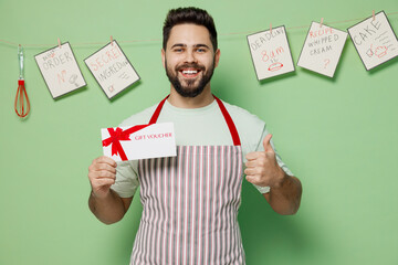 Sticker - Young chef confectioner baker man in striped apron hold store gift certificate coupon voucher card show thumb up isolated on plain pastel light green background studio portrait. Cooking food concept.