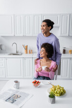 African American Man Hugging Girlfriend With Cup Near Fruits And Newspaper In Kitchen.