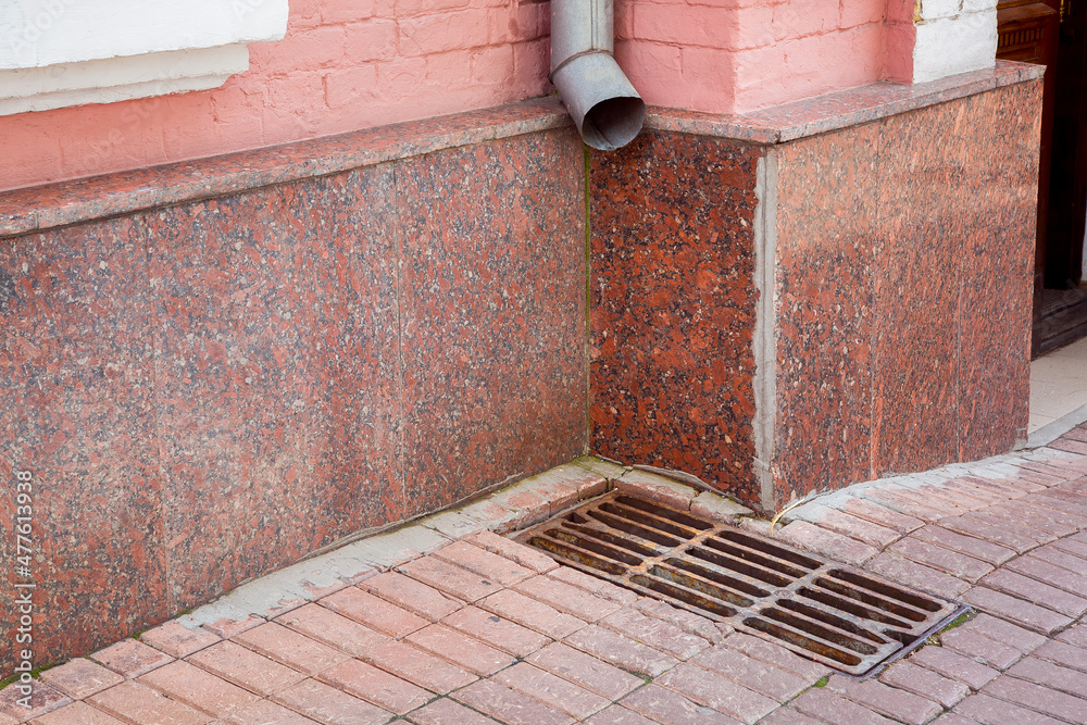 drainage of rainwater a pipe on the wall of the facade of the building with a rain grate of the hatch on the pedestrian sidewalk made of stone tiles near building corner.